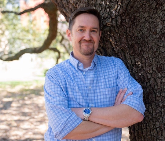 A white man stands in front of a tree and smiles