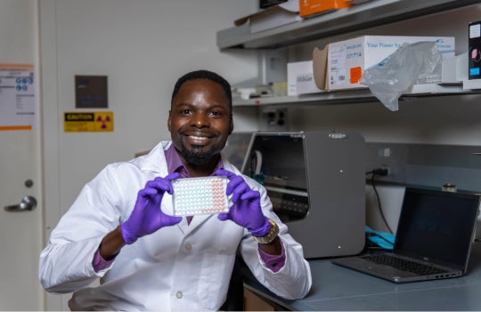 A black man in a lab coat holds a 96 well plate and smiles.