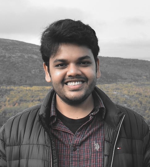 An Indian man stands outside, smiling at the camera. In the distance behind him, there is a large hill.