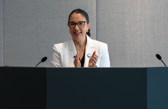 Dr. Karen Lozano, wearing dark-rimmed glasses and a white blazer, smiles and applauds while standing behind a dark podium during a presentation.