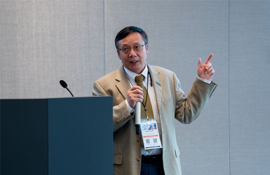 A speaker wearing glasses, a beige suit jacket, a patterned tie, and a conference badge stands behind a podium, holding a microphone in one hand and gesturing upward with his other hand while presenting.
