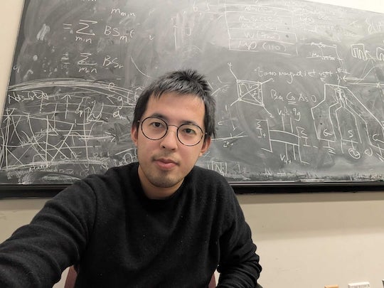 A young Asian man sits in front of a chalkboard filled with equations