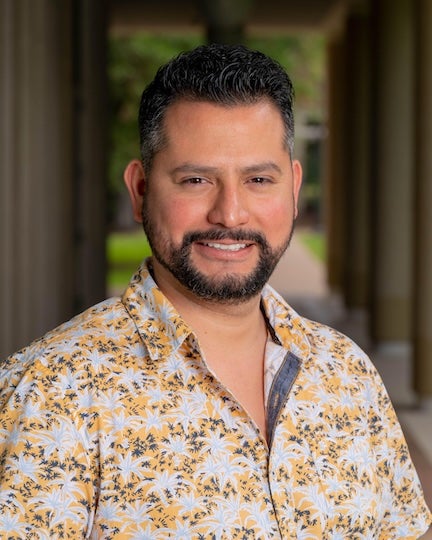 A bearded Latino man in a patterned shirt smiles at the camera. 