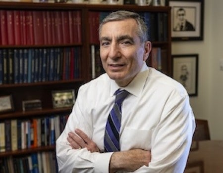 An older white man, standing with arms crossed, is in front of a bookshelf. 