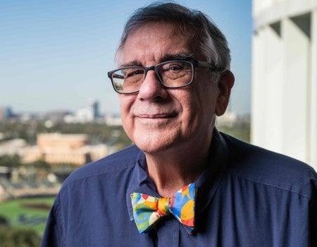 An older white man, Peter Wolynes, smiles slightly in front of a sunny window. He is wearing glasses and a multicolored bowtie.