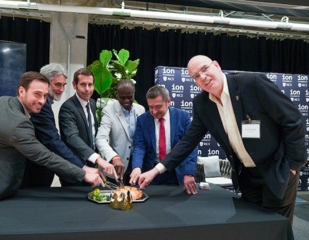Six men, all in suits, are gathered around a cake. Each man is cutting a piece of a cake with a small plastic knife and smiling at the camera