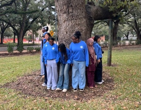 A group of students, including Black, white, and Muslim students stand circled around a tree, with their back to its bark. They are looking at each other or down at the ground.