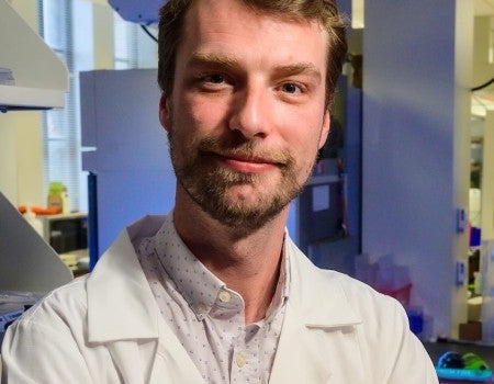 A white man stands in a lab and smiles slightly at the camera