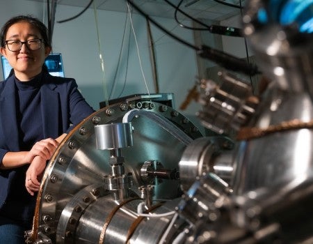An Asian woman, smiling, stands next to a large machine with a bell opening 