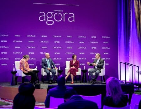 A group of people sit on a stage, having a discussion in front of a purple backdrop