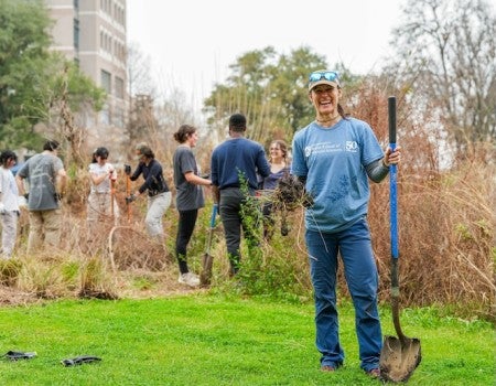 A white woman with a shovel stands smiling in a prairie. A group of people are working on the prairie behind her. In the distance is a large commercial building