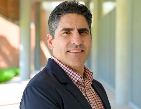 A man in a suit stands in an outdoor hallway, smiling slightly