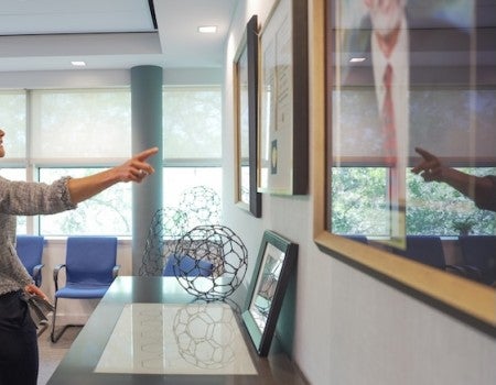 A brunette white woman, the Crown Princess of Sweden, stands pointing at a framed medal on a wall. 