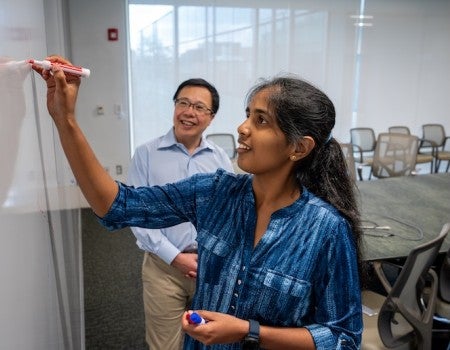 A smiling South Asian woman writes on a whiteboard. Behind her, a smiling East Asian man looks at what she is writing. 