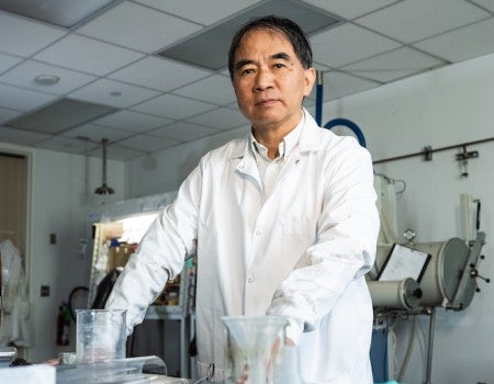 An older Asian man in a lab coat stands at a lab bench. 