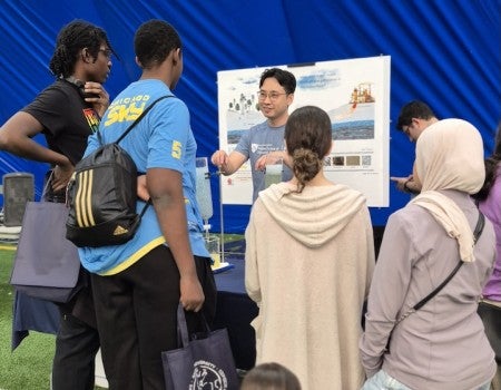 A male Asian graduate student stands in front of a poster, explaining science to a diverse group of kids