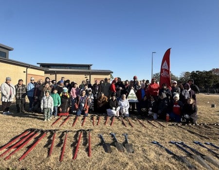 A group of people standing outside on a sunny day. A row of shovels and tools is laid out on the ground in front of them.