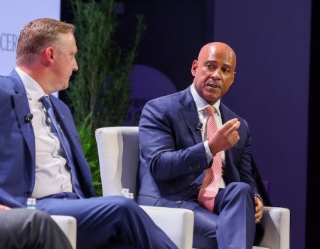 A white man, wearing a suit and sitting on a chair, listens as a Black man, wearing a suit and sitting on the chair next to him, talks.