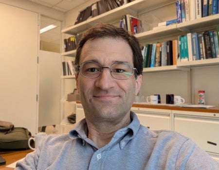 A white man smiles at the camera. He is sitting in front of a wall of books and wearing glasses.