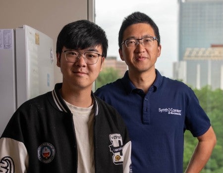 A young Asian man in a letterman jacket stands in front of a middle-aged Asian man in a polo shirt. Both are wearing glasses.