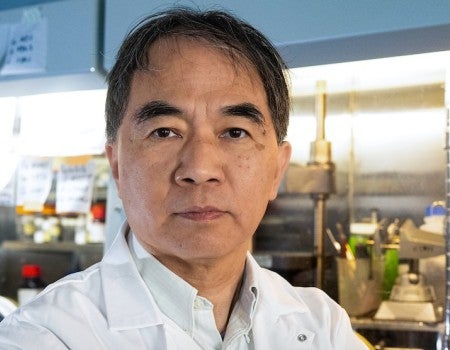 An Asian man in a lab coat stares confidently at the camera. 