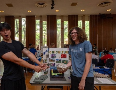 An Asian man excitedly points at a mushroom growth brick held by a laughing white man with curly hair