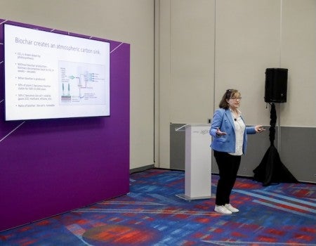 A white woman stands in front of a podium, presenting a slide deck projected on the wall behind her
