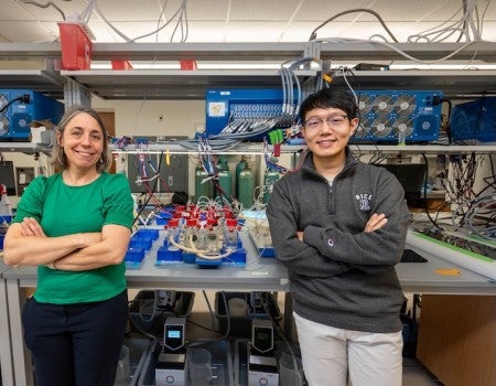 An older white woman and a young Asian man stand in front a lab bench with bottles and wires on it. They are smiling at the camera