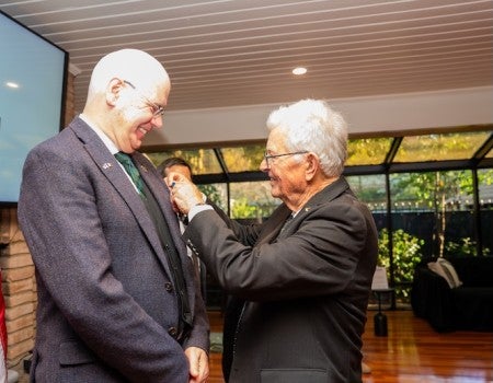 A tall white man stands and smiles at a shorter white man pinning something on his suit coat.