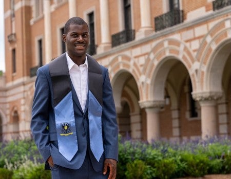 A Black man in a suit and blue stole stands in a university courtyard.