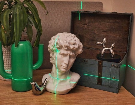 photo of objects with different reflectivity, including a gypsum bust, some shiny metallic figurines a plant, a green chest and green cactus-shaped vase, with green laser light shining across part of the image 