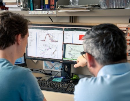 Two white men look at a computer screen which has graphs and datasets on it.