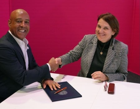 A black man and a white woman, both wearing suits, shake hands and smile