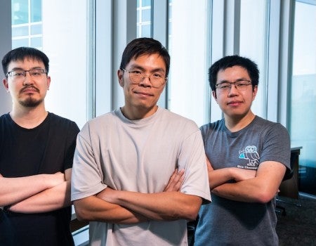 Three Asian men stand with their arms crossed in front of a window.