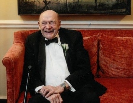 A grinning older white man sits on a red couch. 