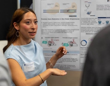 A young woman gestures at a poster while talking to an audience.