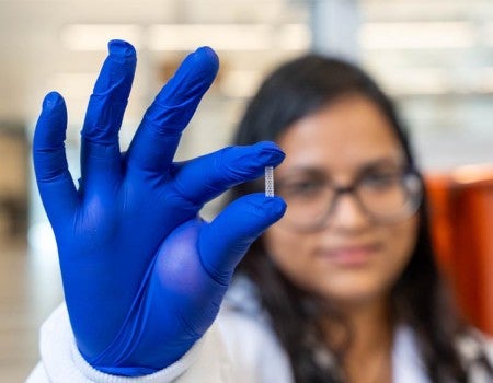 A person in blue gloves holds a tiny, patterned object, with a blurred background showing another individual in a lab setting.