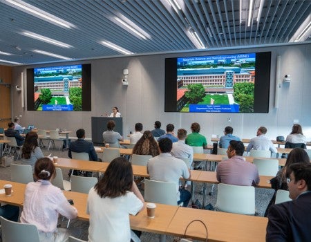 A speaker stands at a podium in a modern university lecture hall, presenting slides titled "Materials Science and NanoEngineering (MSNE)" on two large screens to an audience of university leaders and researchers seated at long wooden tables facing the front.
