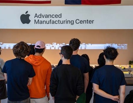 A group of students viewed from behind stands in front of a display at Apple’s Advanced Manufacturing Center, beneath a large wall sign featuring the Apple logo and center name.