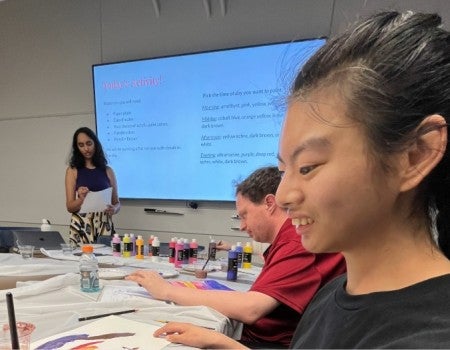A smiling asian woman sits at a table; a woman behind her reads notes in front of a presentation