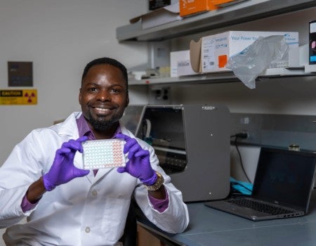 A black man in a lab coat and gloves holds up a 96 well plate while smiling