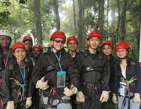 A diverse group of eight young people wearing safety helmets, harnesses, and protective gloves pose together outdoors on a platform surrounded by a lush forest.