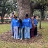 A group of students, including Black, white, and Muslim students stand circled around a tree, with their back to its bark. They are looking at each other or down at the ground.