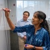 A smiling South Asian woman writes on a whiteboard. Behind her, a smiling East Asian man looks at what she is writing. 
