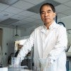 An older Asian man in a lab coat stands at a lab bench. 