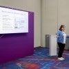 A white woman stands in front of a podium, presenting a slide deck projected on the wall behind her