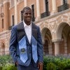 A Black man in a suit and blue stole stands in a university courtyard. 