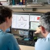 Two white men look at a computer screen which has graphs and datasets on it.