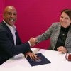 A black man and a white woman, both wearing suits, shake hands and smile 