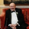 A grinning older white man sits on a red couch.  